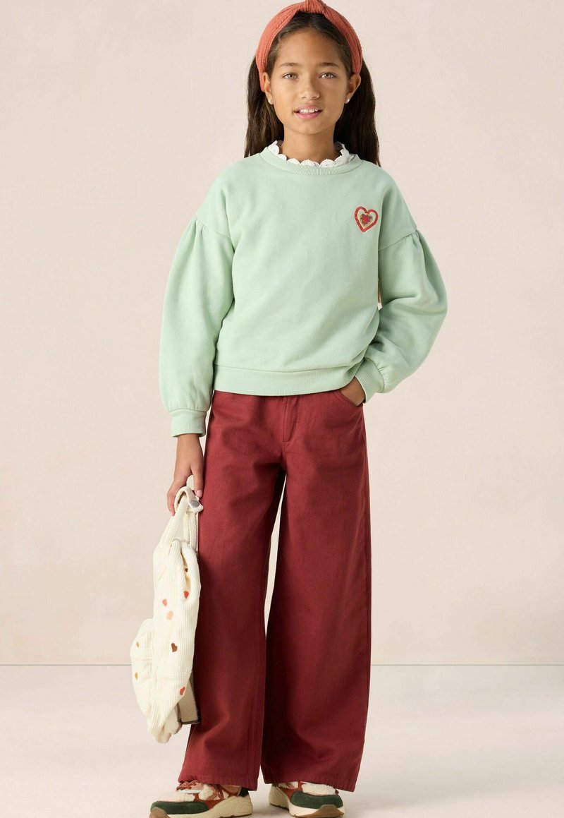 Sweatshirt vert menthe avec des manches bouffantes et une broderie en forme de cœur, associé à un pantalon bordeaux large. Un sac beige avec des accents à motifs.