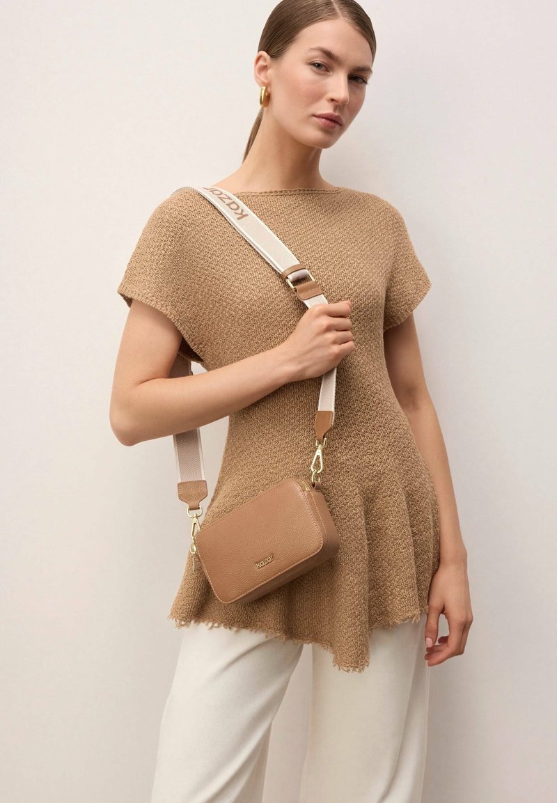 Naine, kes kannab tekstuurse pruunika lühikese varrukaga topi ja valgeid pükse, hoides väikest pruunikat risti üle keha kantavat kotti laia brändi trükiga rihmaga.