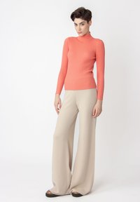 Turtleneck-Pullover aus korallfarbenem Rippstrick, kombiniert mit beigen, weit geschnittenen Hosen aus weichem Stoff, die eine entspannte Silhouette bieten.