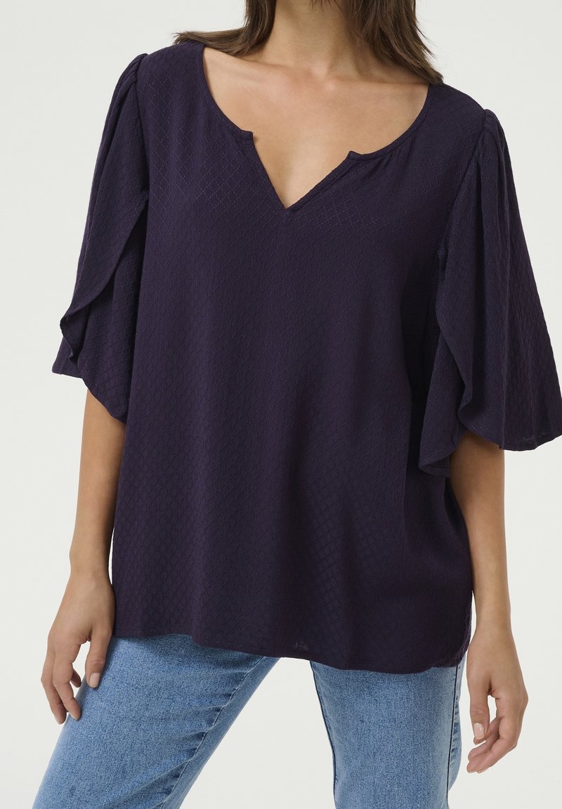 Femme portant un chemisier ample bleu marine foncé en tissu texturé, col en V et manches larges 3/4, associé à un jean bleu clair.