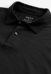 Polo negro hecho de algodón suave, con un cuello clásico, tapeta de dos botones y una sutil textura en la tela. Estilo de corte regular.