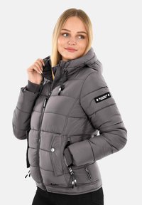 Woman wearing a gray padded hooded jacket with zip and pockets, looking to the side and holding the hood drawstring.
