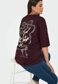 Burgunderfarget oversized t-skjorte med grafisk design på baksiden som viser tekst og et båndillustrasjon. Mykt stoff, avslappet passform, kortermet.