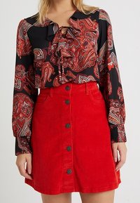 Svart blus med röd paisleymönster, volangkrage och en röd kord-flodkjol med knappar framtill och svarta knappar.