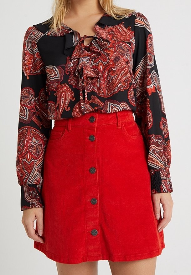 Svart blus med röd paisleymönster, volangkrage och en röd kord-flodkjol med knappar framtill och svarta knappar.