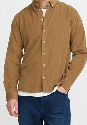 Mand iført en brun kordfløjls-skjorte med knapper over en hvid undertrøje, med mørkeblå jeans, stående med den ene hånd i lommen.