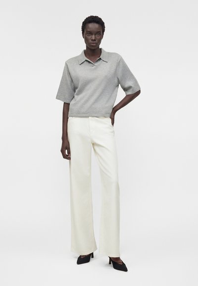 Γυναίκα στέκεται με το ένα χέρι στη μέση, φοράει γκρι κοντομάνικη μπλούζα με γιακά, άσπρο παντελόνι με φαρδιές μπατζάκια και μαύρα μυτερά τακούνια.