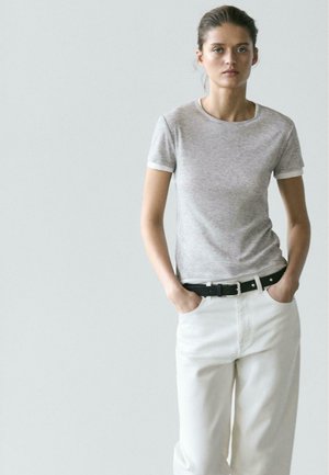 Femme debout, les mains dans les poches, portant un t-shirt gris à manches courtes, un pantalon blanc et une ceinture noire, sur un fond uni.