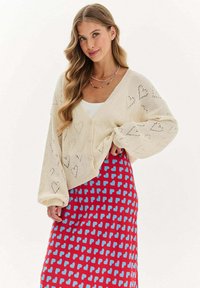 Krémový pletený kardigán s vzorom srdca, výstrih do V a gombíkmi. V kombinácii s červenou sukňou so vzorom modrých sŕdc. Hladká textúra.