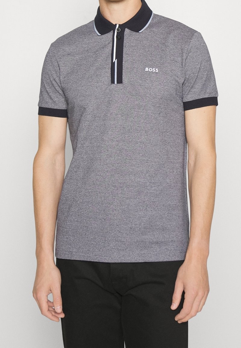 Homme portant un polo gris à manches courtes avec col et bordure des manches noirs, et le logo "BOSS" sur la poitrine gauche, associé à un pantalon noir.