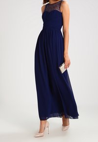 Navy blue maxi dress with pleated bodice, sheer lace neckline, and flowing skirt. Paired with nude high heels and a small metallic clutch.