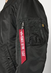 Schwarze Bomberjacke mit einer Reißverschlusstasche am linken Ärmel, rotem "REMOVE BEFORE FLIGHT"-Tag und strukturiertem, glattem Stoff.