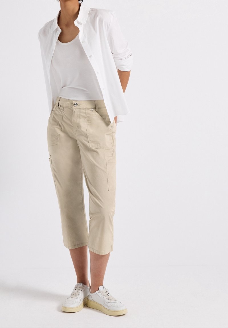 Kvinna som bär beige, korta cargo-byxor, vit linneöverdel, vit, uppknäppt skjorta och vita sneakers, stående med händerna i fickorna.