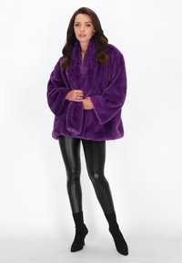 Femeie îmbrăcată într-o jachetă din blană artificială purpurie, leggings din piele negre și bocanci cu toc negri, stând în fața unui fundal alb.