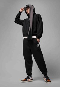 Zwarte cropped hoodie met een voorzak, gecombineerd met zwarte sweatpants met een logo-embleem. De schoenen zijn zwart met oranje sneakers.
