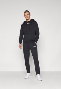 Set di felpa e pantaloni nera realizzato in tessuto morbido. La felpa presenta i lacci e una tasca frontale; i pantaloni hanno tasche laterali e un logo bianco.