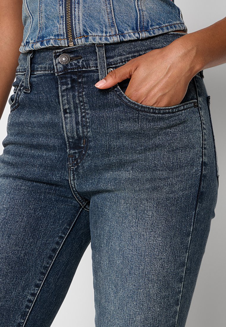 Main gauche d'une femme reposant dans la poche avant d'un jean en denim taille haute de couleur bleu foncé, avec des détails de bouton et de fermeture éclair visibles.