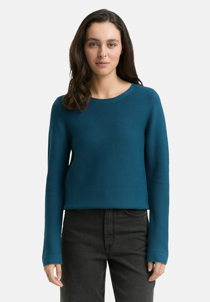 Türkiser gerippter Pullover mit langen Ärmeln und Rundhalsausschnitt. Die Stofftextur wirkt weich und dehnbar, kombiniert mit schwarzer High-Waist-Hose.
