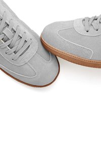 Graue Wildledersneaker mit strukturiertem Obermaterial, flachen grauen Schnürsenkeln und brauner Gummisohle mit einem gemusterten Rand.