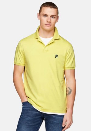 Camisa polo amarilla de algodón. Presenta un cierre de dos botones y un pequeño logo bordado en el lado izquierdo del pecho. Se lleva con jeans azul.