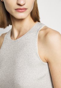 Close-up van het bovenste deel van de torso van een vrouw die een mouwloze, gestructureerde zilveren top met hoge hals draagt, waarbij een deel van haar lippen en schouder zichtbaar is.