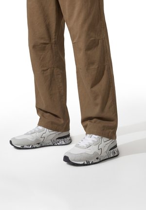 Weiße und graue Freizeitsneaker mit schwarzen, gemusterten Sohlen, getragen zu lockeren braunen Hosen vor weißem Hintergrund.