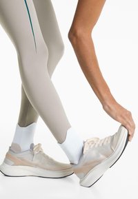 Egy ember bézs leggingset és fehér zoknit viselve a lábát nyújtja, miközben a lábfejét egy fehér és bézs futócipőben tartja, fehér háttér előtt.