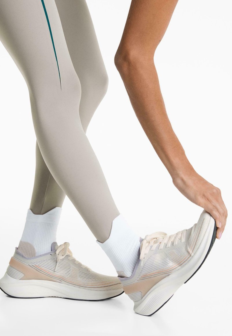 Egy ember bézs leggingset és fehér zoknit viselve a lábát nyújtja, miközben a lábfejét egy fehér és bézs futócipőben tartja, fehér háttér előtt.