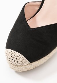 Espadrilles en suedine noire avec un capuchon en crochet beige et une semelle en jute. Le design présente une découpe en forme de V à l'avant.