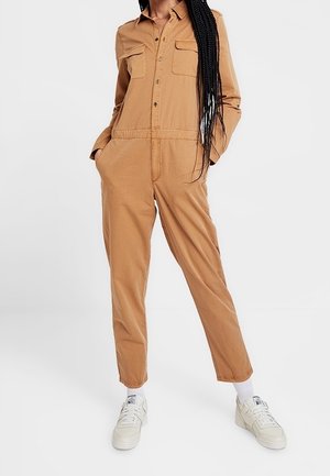 Kvinde iført en beige knap-op-dragt med forlommer, hænderne i lommerne, hvide sneakers og langt fletning hår lagt over skulderen.