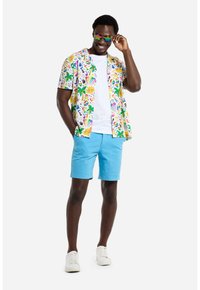 Camicia a maniche corte con bottoni e una stampa tropicale colorata, abbinata a pantaloni corti azzurri e sneakers bianche. Accompagnata da occhiali da sole multicolori.