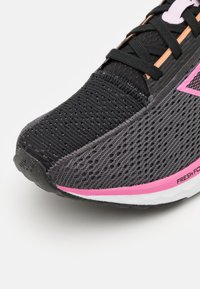 Zapatilla deportiva negra con parte superior de malla, detalles texturizados, franja de acento rosa y suela blanca. Presenta materiales transpirables y un sistema de cordones resistente.