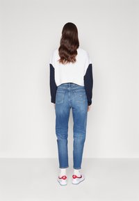 Suéter blanco acanalado con mangas azul marino, combinado con jeans de mezclilla azul claros y zapatillas blancas con acentos rojos. Vista trasera con cabello de longitud media.