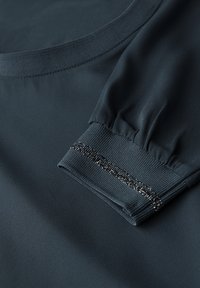 Mörkt tångfärgad blus med rundad halsringning, rynkade ärmar och elastiska manschetter som har en metalltråd detaljer längs manschetten. Slät textur.