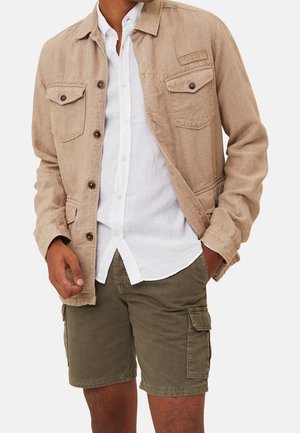 Homme portant une veste beige à boutons par-dessus une chemise blanche, avec un short cargo vert olive, mains dans les poches, sur un fond uni.
