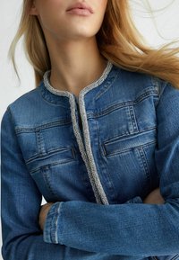 Mladá žena s dlouhými blond vlasy, oblečená v modré džínsové bundě s kamínkovou ozdobou kolem límce a předního zapínání.