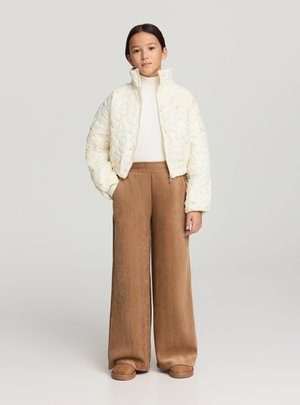 Giacca bomber imbottita crema con motivo floreale, dolcevita bianco e pantaloni marroni a gamba larga. La modella posa su uno sfondo neutro.