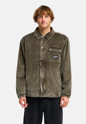 Young man with light brown hair wearing a brown corduroy jacket with snap buttons and black pants, standing against a plain background.