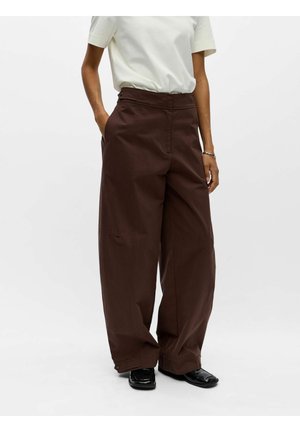 Personne portant un pantalon marron à jambes larges, une chemise blanche à manches courtes et des chaussures noires à bout carré, debout avec une main dans la poche.
