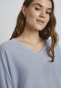 Pulover bleu deschis cu decolteu în formă de V, realizat dintr-un material texturat, cu o croială lejeră și umeri ușor lăsați. Cercei circulare aurii vizibili.
