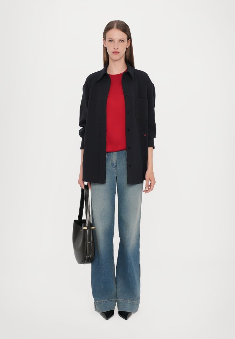 Chemise boutonnière bleu marine sur un haut rouge, associée à un jean flare bleu et des escarpins noirs à bout pointu. Tient un sac à main noir avec des accents dorés.
