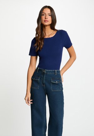 Frau mit langen braunen Haaren, die ein marineblaues, geripptes Kurzarmshirt und hoch taillierte dunkelblaue Jeans mit Frontklappen-Taschen trägt.