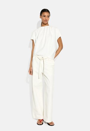 Kobieta ubrana w luźną białą bluzkę z krótkim rękawem, białe spodnie z wysokim stanem i szerokimi nogawkami ze sznurkiem oraz czarne sandały z czerwonymi paskami.