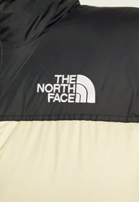 Zwarte en beige jas met een hoge kraag; heeft het logo van "The North Face" in witte borduring op de borst. Soepele stofstructuur.
