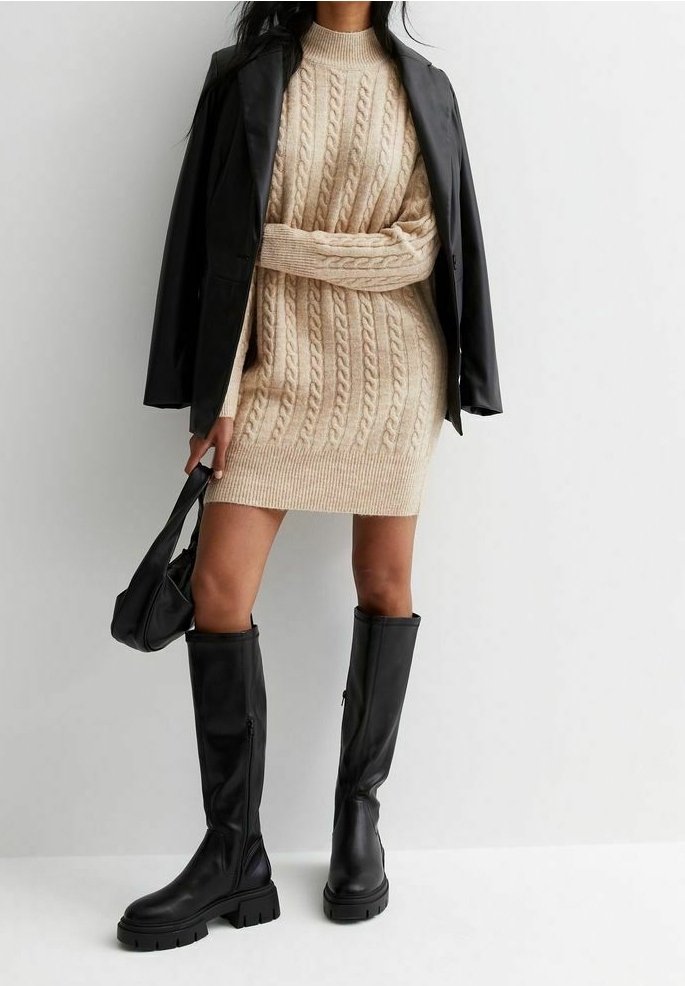 Femme portant une robe-pull beige en maille torsadée, blouson en cuir noir posé sur les épaules, bottes noires montantes et tenant un sac à main noir.
