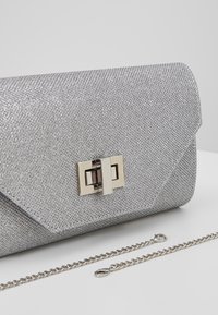 Sølvmetallic clutch taske med tekstureret overflade, vinkelret klap og et guld hardwarelås. Har en aftagelig kædestrop.