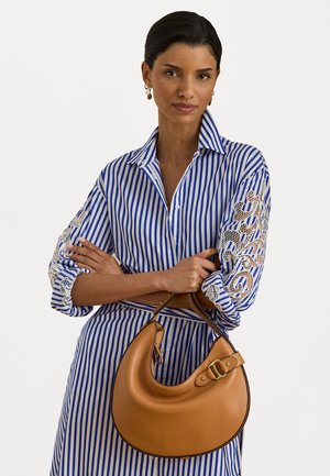 Tan leather hobo bag with a curved shape, gold hardware accent, and a detachable strap. Model wears a blue and white striped shirt with lace details.