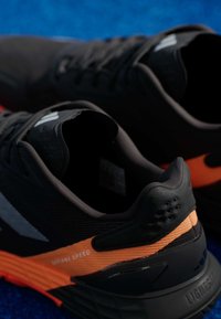 Αθλητικά παπούτσια με μαύρο συνθετικό επάνω μέρος, υφασμάτινη υφή, πορτοκαλί ελαστικές λεπτομέρειες και μαξιλαρένια σόλα με κομψό σχέδιο.