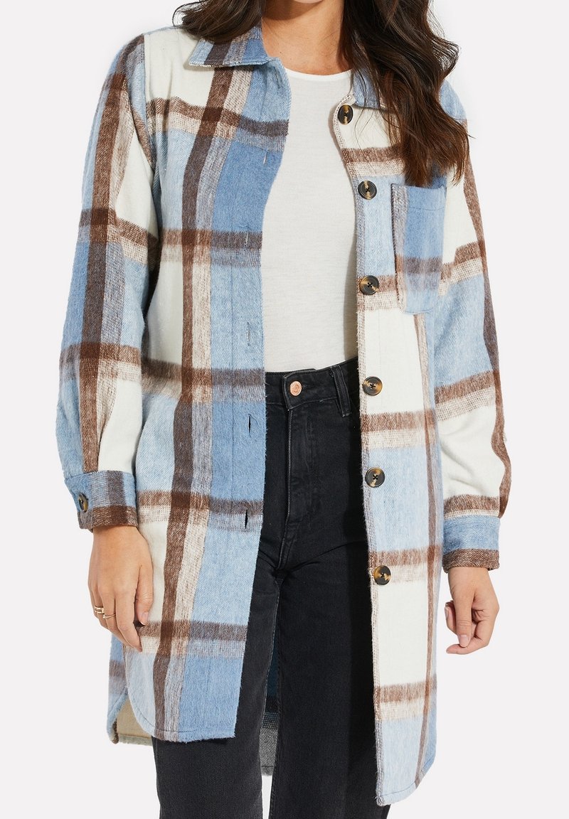 Femme portant une longue veste à carreaux bleu, marron et blanc, boutonnée, par-dessus un top blanc et un jean noir, debout devant un fond uni.
