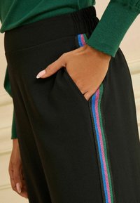 Zwarte broek met een gestructureerde tailleband, voorzien van een strak zijzakje versierd met blauwe, groene en roze metallic strepen.